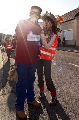 Ein Mann und eine Frau in Kostümen und Warnwesten stehen mitten auf der Straße, lächeln und posieren für ein Foto. Hinter ihnen gehen Menschen in Warnwesten die Straße entlang.