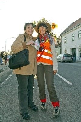 Zwei Frauen stehen auf der Straße, eine trägt eine Warnweste und einen Hut mit Sonnenblumen, die andere eine Handtasche. Sie lächeln beide und scheinen für ein Foto zu posieren. Hinter ihnen ist ein Gebäude und ein geparktes Auto zu sehen.