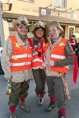 Drei Personen in Militärkostümen mit reflektierenden orangefarbenen Westen und Hüten stehen vor einem Gebäude mit einem Schild, das 'Cafe Salotti' besagt.