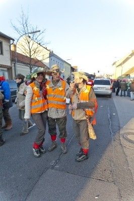 Drei Personen in orangefarbenen Sicherheitswesten und Hüten stehen in der Mitte einer Straße, mit mehreren Personen und Fahrzeugen im Hintergrund.