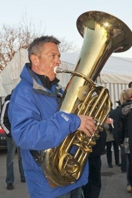 Ein Mann in einer blauen Jacke spielt ein großes Messing-Tuba im Freien, mit mehreren Personen im Hintergrund.