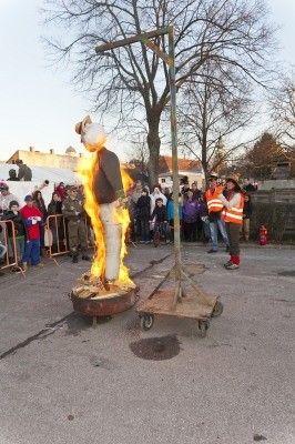 Eine brennende Strohpuppe auf einem Wagen bei einer Outdoor-Veranstaltung mit Zuschauern und einem Feuerlöscher in der Nähe.