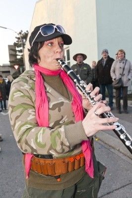 Eine Frau in Tarnkleidung und einem rosa Schal spielt Klarinette im Freien, während mehrere Personen hinter ihr stehen.