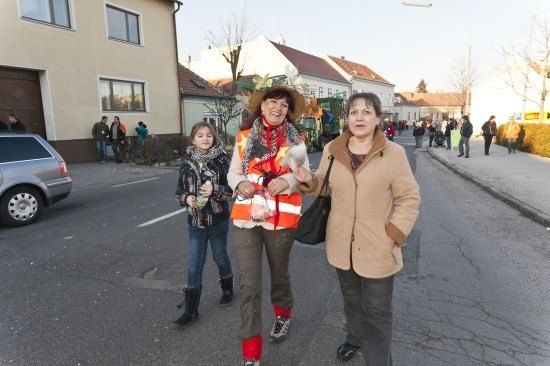 Drei Frauen gehen auf einer Straße mit Gebäuden und geparkten Autos im Hintergrund, eine trägt eine reflektierende Weste und einen Hut.