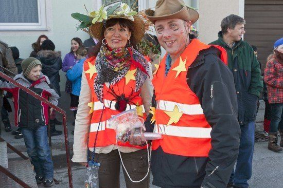 Zwei Personen in orangenen Westen mit gelben Sternen, eine trägt einen Strohhut mit Blumen, posieren für ein Foto mit einer Menschenmenge im Hintergrund.