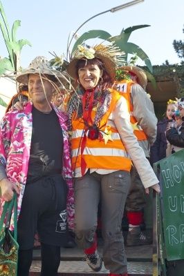 Zwei Menschen in bunten Kostümen und Hüten stehen auf einer Bühne, wahrscheinlich bei einem Festival. Der Mann links trägt eine pinke Jacke und schwarze Hose, während die Frau rechts eine orange Weste und graue Hose trägt. Hinter ihnen stehen Menschen in Kostümen und ein grünes Banner mit weißem Text.