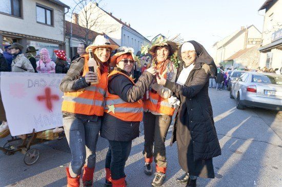 Vier Frauen in orangefarbenen Westen und Hüten posieren für ein Foto auf einer Straße, mit einem Banner und einem Schubkarren im Hintergrund.