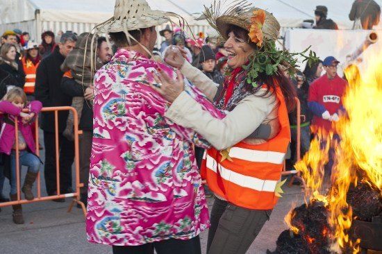 Zwei Frauen tanzen vor einem Lagerfeuer, eine trägt ein Blumenhemd und die andere eine orange Weste mit Grünzeug auf dem Hut. Eine Menge beobachtet hinter einer Barriere.