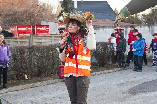 Eine Frau in einer Warnweste und einem mit Blumen verzierten Strohhut spricht in ein Mikrofon auf einer Straße. Hinter ihr versammeln sich Kinder und Erwachsene, einige in roten Jacken.