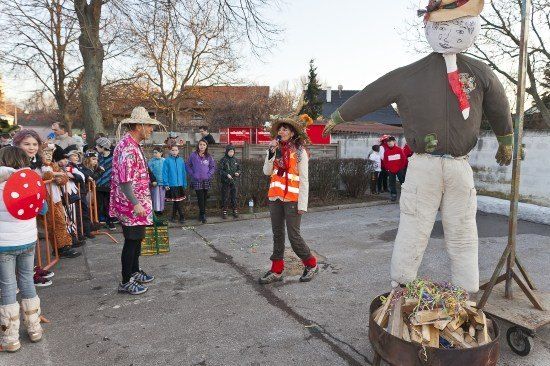 Eine Gruppe von Menschen, einschließlich Kinder, versammelt sich um eine große Strohfigur, möglicherweise für ein Fest. Eine Frau in einer orangefarbenen Weste hält ein Mikrofon.