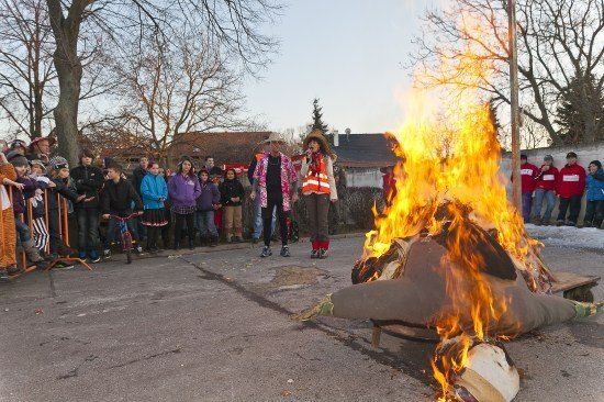 Eine Menschenmenge beobachtet, wie eine Person mit Hut und Weste in ein Mikrofon singt, in der Nähe einer brennenden Puppe auf der Straße.