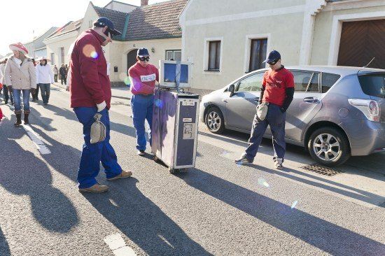 Zwei Männer mit einem Wagen gehen auf der Straße. Ein silberner Wagen ist auf der Seite geparkt. Menschen gehen in die entgegengesetzte Richtung.