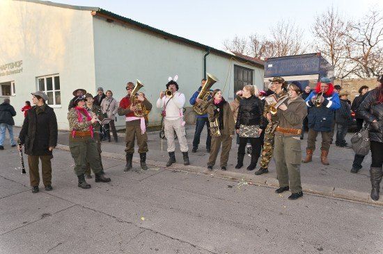 Eine Gruppe von Menschen in militärischen Uniformen und Mützen steht auf der Straße und spielt Musikinstrumente. Ein Gebäude mit einem Schild steht hinter ihnen.