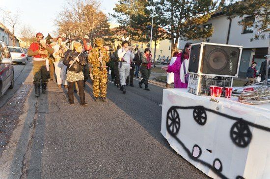 Eine Gruppe von Menschen in verschiedenen Kostümen, darunter Militär- und Tieroutfits, marschiert eine Straße entlang, in der Nähe eines dekorierten Tisches und eines Lautsprechers.