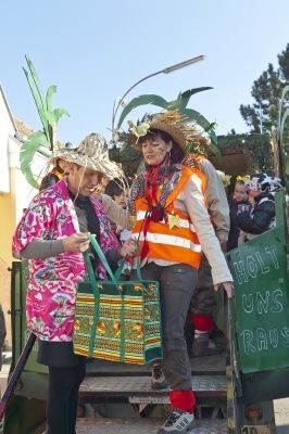 Zwei Frauen in Hüten und Westen, eine hält eine farbenfrohe Tasche, stehen auf einer Plattform mit einem Schild, das 'Holy Uns Raus' steht. Sie scheinen an einem Umzug oder Festival teilzunehmen.