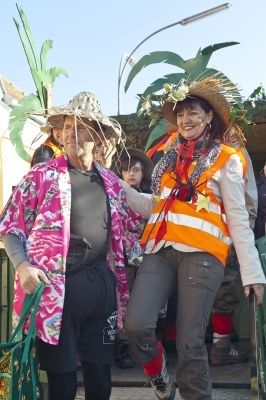 Zwei Personen in Kostümen, eine in einer rosafarbenen Blumenjacke und Hut, die andere in einer orangefarbenen Weste und Hut, stehen nebeneinander. Sie scheinen an einer Parade teilzunehmen.
