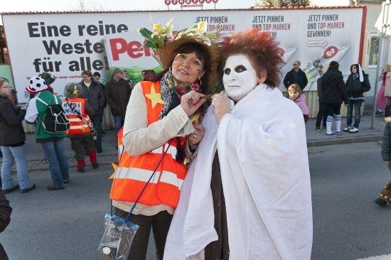 Eine Frau in einer orangefarbenen Weste und ein Mann in einem weißen Umhang mit rotem Haar posieren für ein Foto auf einer Straße mit einem großen Schild im Hintergrund.