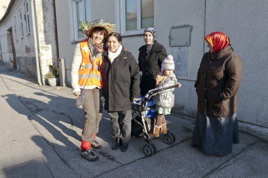 Fünf Personen, darunter eine Frau mit einem Baby im Kinderwagen, stehen auf einer Straße vor einem Gebäude.
