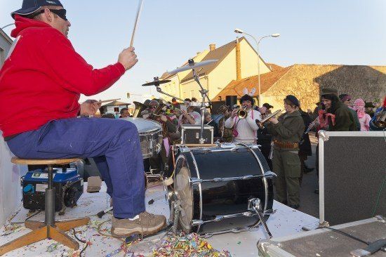 Ein Mann in einer roten Kapuzenjacke spielt auf einer Bühne Schlagzeug mit einer Brassband im Hintergrund.