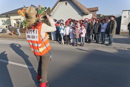 Eine Person in einer Warnweste und Hut fotografiert eine Gruppe von Kindern in roten Hüten, die vor einem Haus stehen.