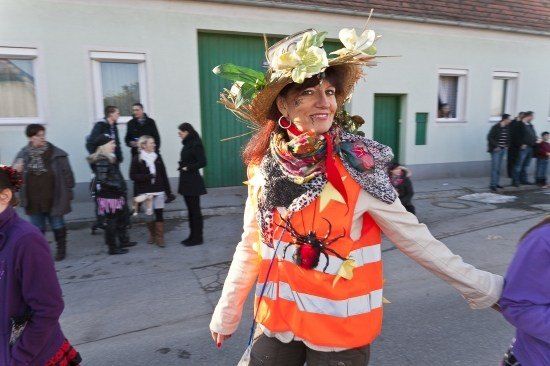 Eine Frau in einer Warnweste und einem Hut mit Blumen geht eine Straße entlang, mit anderen Leuten im Hintergrund.