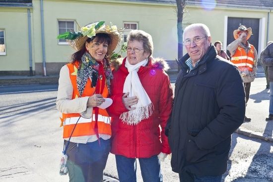 Drei Menschen stehen an einem sonnigen Tag draußen. Eine Frau links trägt einen Strohhut mit Blumen, eine andere Frau in der Mitte trägt einen roten Mantel und einen weißen Schal, und ein Mann rechts trägt einen schwarzen Mantel. Hinter ihnen ist ein Gebäude mit Fenstern und einem Baum zu sehen.
