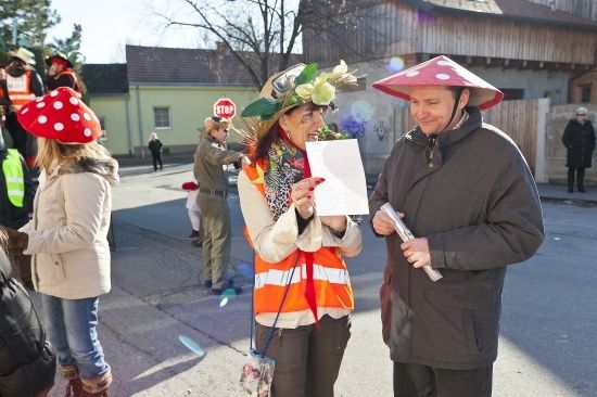 Eine Frau in einer Warnweste und einem Pilzhut liest ein Stück Papier, während ein Mann in einem Pilzhut neben ihr lächelt auf einer Straße.