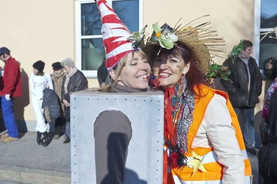 Zwei Frauen in Kostümen, eine mit einer Metallkastenmütze und die andere mit einem Strohhut mit Blumen, lächeln für die Kamera. Hinter ihnen gehen Leute auf einem Bürgersteig.