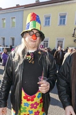 Eine Person in einem bunten Clown-Kostüm mit einer großen roten Nase, die einen Becher hält, steht vor einem Gebäude mit mehreren Personen im Hintergrund.