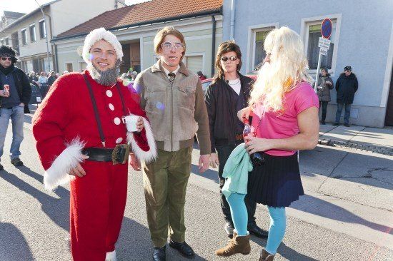 Eine Gruppe von Menschen in verschiedenen Kostümen, darunter ein Weihnachtsmann-Kostüm, steht auf einer Straße mit Gebäuden im Hintergrund.