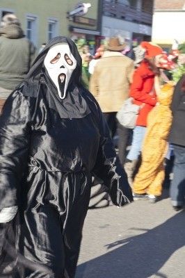 Eine Person in einem schwarzen Kostüm mit einer weißen Maske und einem Schrei-Gesicht läuft auf der Straße mit anderen Leuten während eines Umzugs.