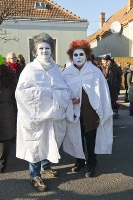 Zwei Personen in weißen Roben und Gesichtsbemalung, möglicherweise bei einem Umzug oder Festival, stehen zusammen auf einer Straße mit anderen Menschen im Hintergrund.