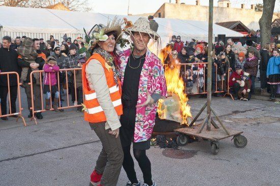 Ein Mann und eine Frau stehen vor einer brennenden Puppe, umgeben von einer Menschenmenge, einige in Kostümen, die zuschauen.