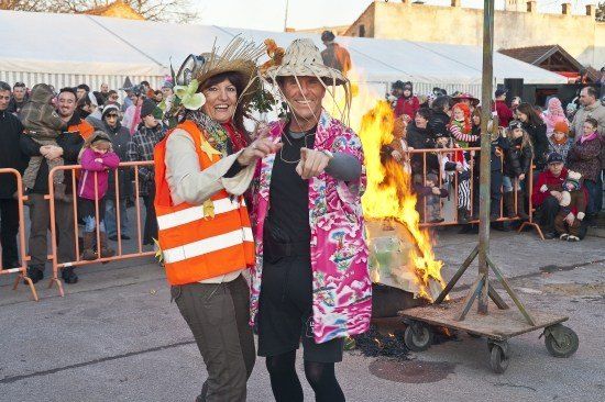 Zwei Menschen in Kostümen stehen neben einem brennenden Scheiterhaufen bei einer Outdoor-Veranstaltung, die von einer Menge beobachtet wird.