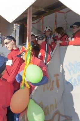 Eine Gruppe von Menschen in roten Hemden und Mützen steht in einem dekorierten Bereich, hält Luftballons und posiert für ein Foto. Die Wand hinter ihnen hat Graffiti und ein Schild, das 'Bank' besagt.