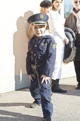 Ein junger Junge in einer Polizeiuniform, der auf einem Bürgersteig steht, mit einer Frau in einem weißen Kleid und einer anderen Frau in einem braunen Mantel hinter ihm.