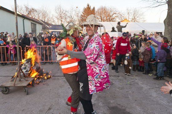 Zwei Menschen in bunten Kostümen tanzen vor einer Menge in der Nähe eines brennenden Feuers.
