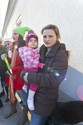 Eine Frau hält ein junges Kind in einer pink-weiß karierten Jacke, während sie neben einer anderen Frau in einem roten Mantel steht, beide blicken in einem winterlichen Umfeld nach vorne.
