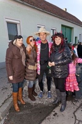 Fünf Personen stehen vor einem Gebäude, drei Frauen in Mänteln und Stiefeln, ein Mann in Hut und Sonnenbrille und ein Kind in einem rosa Kleid.