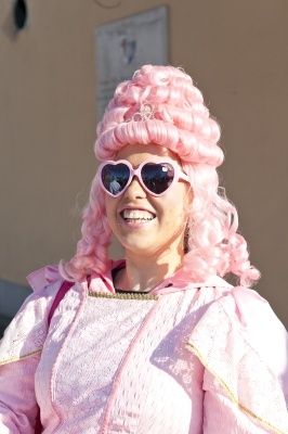 Eine Frau mit rosa lockigem Haar, die ein rosa Kostüm und Sonnenbrille mit herzförmigen Gläsern trägt und für die Kamera lächelt.