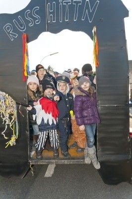 Eine Gruppe von Kindern in verschiedenen Kostümen, darunter ein Pirat und ein Polizist, steht in einem dekorierten Wagen mit den Worten 'RUSS HITTN' oben drauf. Sie nehmen wahrscheinlich an einem Umzug teil.