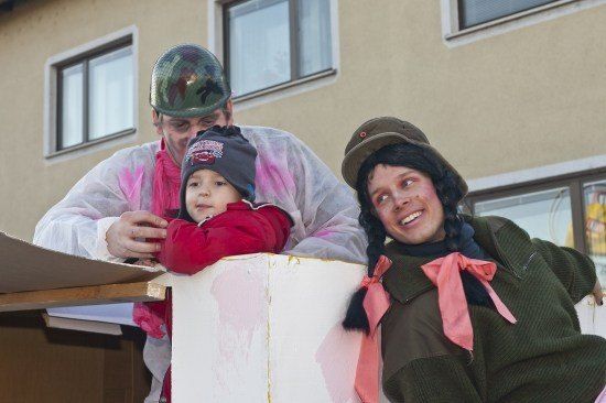 Drei Personen in Militäruniform, einschließlich eines Kindes, stehen vor einem Gebäude, wobei eine das Kind auf einer weißen Struktur hält.