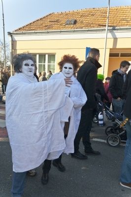Zwei Personen in weißen Umhängen mit Gesichtsbemalung stehen auf der Straße, möglicherweise für eine Kostümveranstaltung, mit einem Kinderwagen und mehreren Personen im Hintergrund.