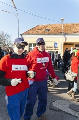 Zwei Männer in roten Hoodies und blauer Hose, mit Masken und Handschuhen, stehen zusammen auf einer Straße, mit einem Gebäude und Menschen im Hintergrund.