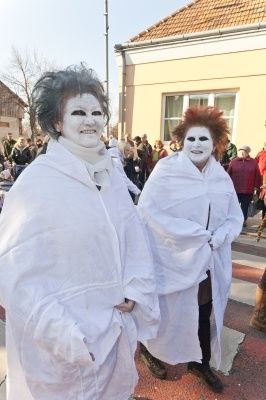 Zwei Personen in weißen Gewändern und Gesichtsbemalung, möglicherweise bei einer Parade oder Veranstaltung, gehen vor einem Gebäude, mit einer Menschenmenge im Hintergrund.