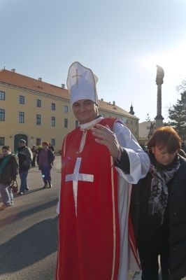 Ein Mann in Bischofskleidung in einem roten und weißen Gewand steht vor einem Gebäude mit einer Frau in einem Schal.