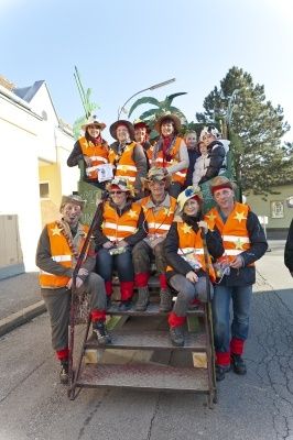Eine Gruppe von Menschen, die orange Westen und Hüte tragen, sitzt auf der Rückseite eines dekorierten Lastwagens und posiert für ein Foto im Freien.