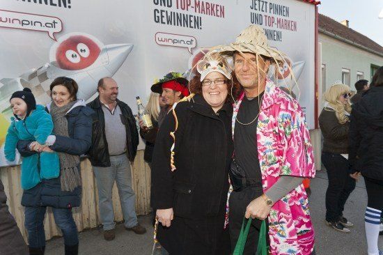 Eine Gruppe von Menschen in Kostümen steht vor einem großen Werbebanner. Das Banner zeigt eine Rakete und Text in einer fremden Sprache. Der Mann und die Frau vorne lächeln und posieren für ein Foto.