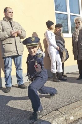 Ein Kind in einer Polizeiuniform führt eine Kampfsportpose vor einem Gebäude aus, während mehrere Erwachsene hinter ihm stehen.