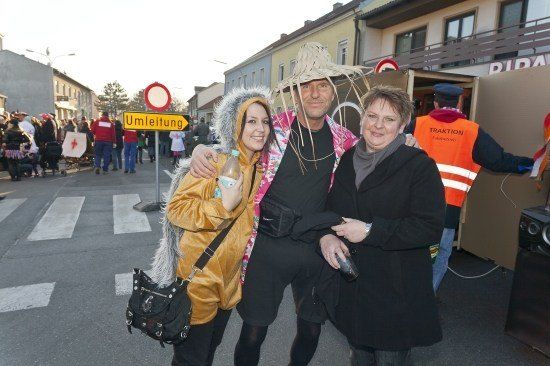 Drei Menschen in Kostümen posieren für ein Foto auf einer Straße mit einer Menschenmenge und einem gelben Schild, das 'Umleitung' steht.